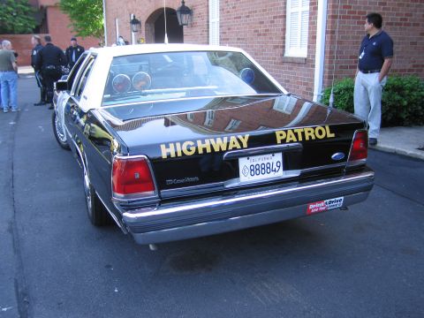 1991 Ford LTD Crown Victoria - glossy black-and-white Highway Patrol sedan photographed from the rear, with people standing near a brick building behind it.