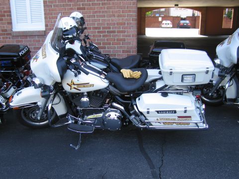 1991 Ford LTD Crown Victoria - white Sheriff Harley-Davidson police motorcycle with side cases and gloves on the seat, parked against a brick wall.