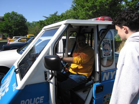 1991 Ford LTD Crown Victoria - man in a yellow shirt sits at the wheel of a blue NYPD three-wheeled police scooter while a boy looks on.