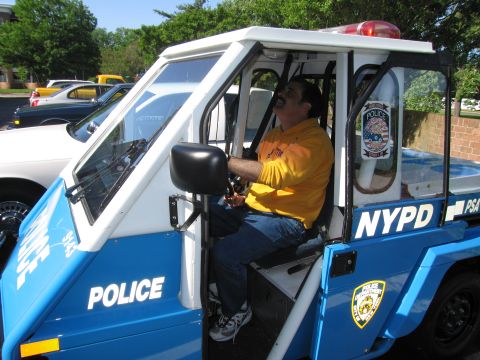 1991 Ford LTD Crown Victoria - mustached man in a yellow shirt sits inside a blue-and-white NYPD three-wheeled police scooter, smiling.
