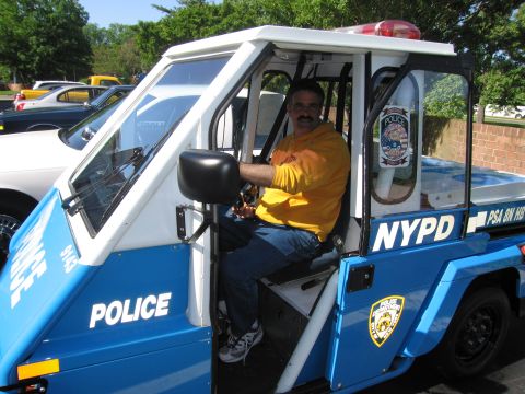 1991 Ford LTD Crown Victoria - man in a yellow shirt sits in a blue-and-white NYPD three-wheeled police scooter at an outdoor gathering.