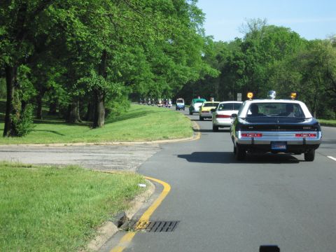 1991 Ford LTD Crown Victoria - police car with roof lights leads a procession of cars and motorcycles down a curving, tree-lined road.