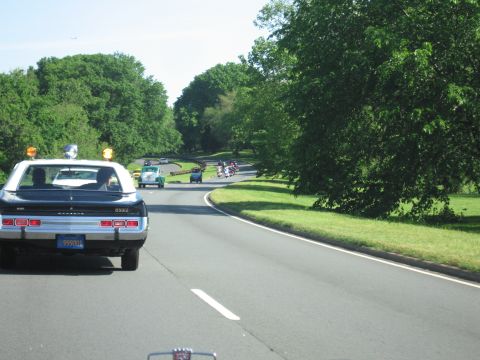 1991 Ford LTD Crown Victoria - black-and-white police car with amber roof lights leads a column of cars and motorcycles along a wooded parkway.