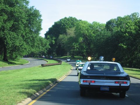 1991 Ford LTD Crown Victoria - black police car with roof beacons leads a convoy of vehicles around a bend on a tree-lined road.