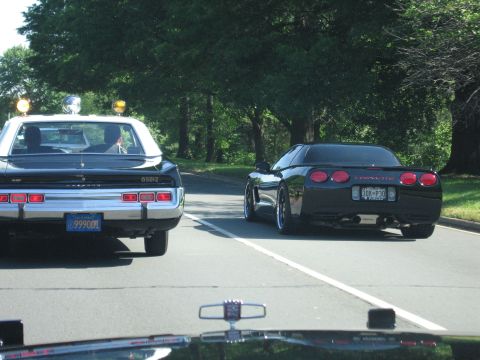 1991 Ford LTD Crown Victoria - black Chevrolet Corvette coupe drives alongside a black police car with roof lights on a tree-lined road.