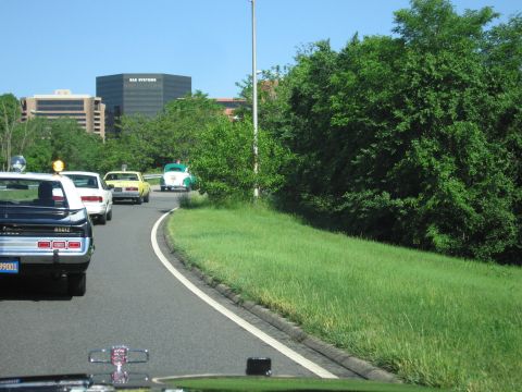 1991 Ford LTD Crown Victoria - black police car and classic cars including a green vehicle travel a parkway with office buildings in the distance.