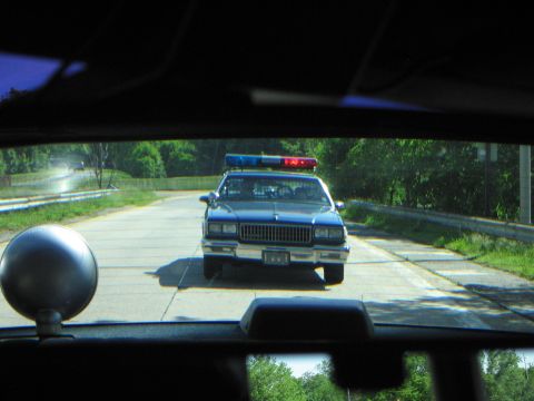 1991 Ford LTD Crown Victoria - View through a windshield of a dark blue police sedan with a roof light bar parked on a paved road lined with trees.