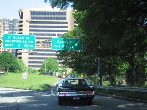 1991 Ford LTD Crown Victoria - dark sedan drives down a tree-lined road beneath green highway signs reading N. Queen St, Courthouse Rd and Rosslyn Key Bridge.