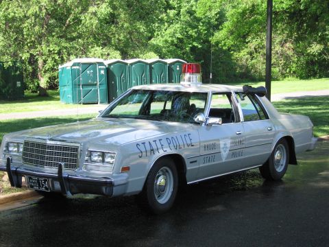 1991 Ford LTD Crown Victoria - silver Rhode Island State Police sedan with a single red roof beacon parked near green portable toilets in a grassy park.