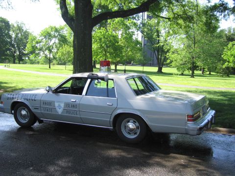 1991 Ford LTD Crown Victoria - Rear three-quarter view of a silver Rhode Island State Police sedan with red roof beacon parked on a wet paved lot in a green park.