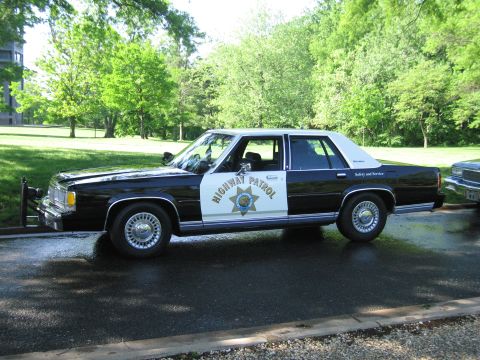 1991 Ford LTD Crown Victoria - Black-and-white Highway Patrol sedan with star shield door marking parked on wet pavement next to another vehicle in a park.
