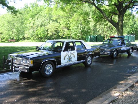 1991 Ford LTD Crown Victoria - black-and-white Highway Patrol sedan parked next to a dark blue police cruiser on a road in a green tree-lined park.