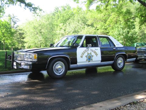 1991 Ford LTD Crown Victoria - Front three-quarter view of a black-and-white Highway Patrol sedan with push bar parked beside another vehicle in a green park.