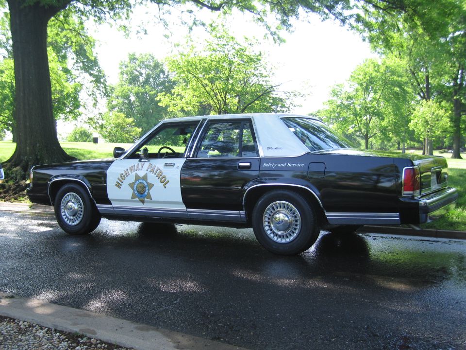 Black and white Highway Patrol sedan in profile under a tree in a park