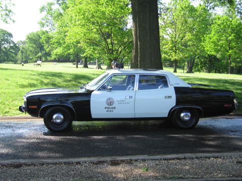 1991 Ford LTD Crown Victoria - Side view of a vintage black-and-white Police sedan with a city shield emblem and unit number, parked on a road in a green park.