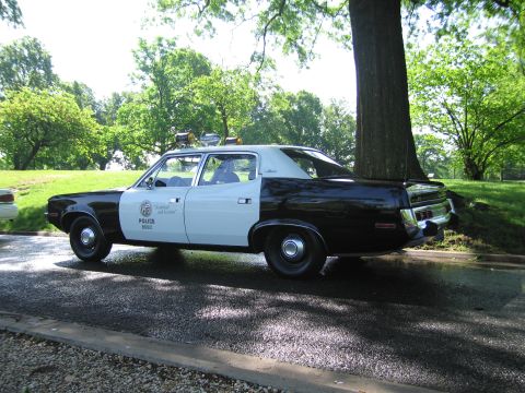 1991 Ford LTD Crown Victoria - Rear three-quarter view of a vintage black-and-white Police sedan with roof lights parked under a tree on wet pavement in a park.