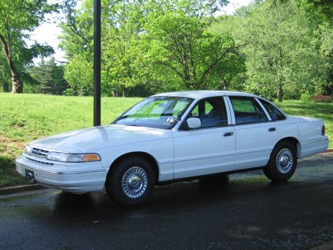 1991 Ford LTD Crown Victoria - Side profile of a white Ford four-door sedan with full wheel covers parked on wet pavement in a sunlit green park.