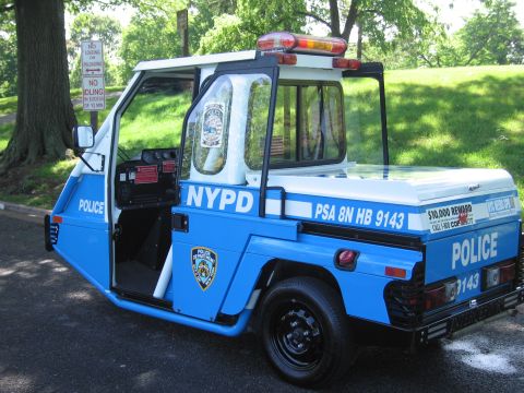 1991 Ford LTD Crown Victoria - blue and white NYPD three-wheeled enclosed police scooter marked PSA 8N HB 9143 parked on pavement near park signage.
