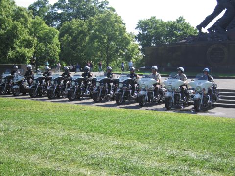 1991 Ford LTD Crown Victoria - row of police motorcycles with helmeted riders lined up on pavement in front of a large bronze figural memorial and trees.