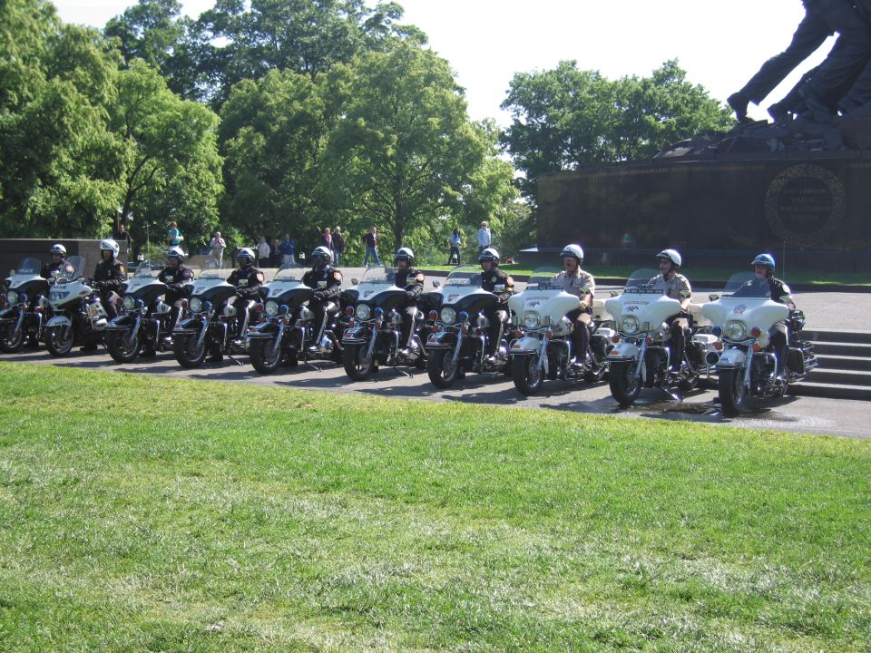 Row of police motorcycles with riders lined up near a bronze memorial statue
