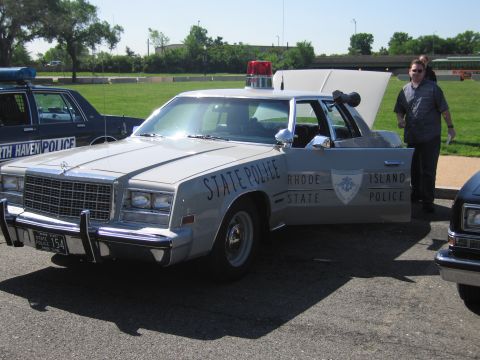 1991 Ford LTD Crown Victoria - silver Rhode Island State Police sedan with a red roof beacon sits in a lot, a man standing beside its open trunk.