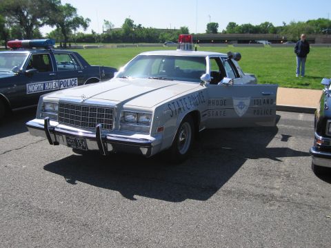 1991 Ford LTD Crown Victoria - silver Rhode Island State Police sedan shown front three-quarter, with chrome bumper, push bar and roof beacon in a grassy lot.