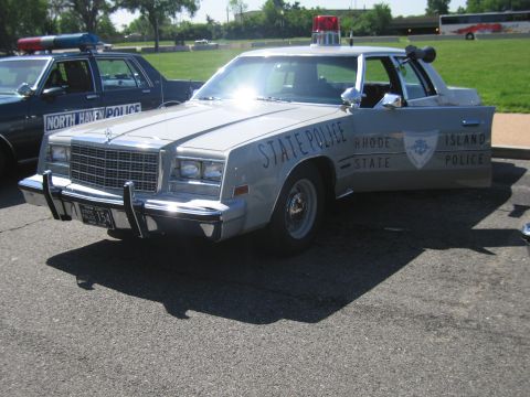 1991 Ford LTD Crown Victoria - silver Rhode Island State Police sedan with a red roof beacon and chrome push bar, sunlight glaring off its hood.