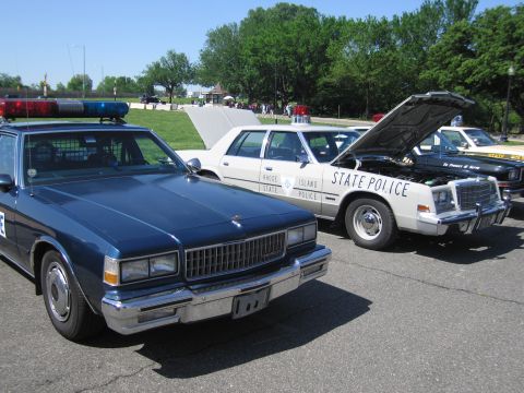 1991 Ford LTD Crown Victoria - row of vintage police cars in a lot, including a dark blue sedan in front and white State Police cars with one hood raised.