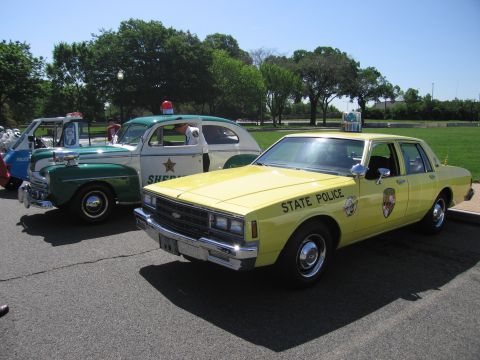 1991 Ford LTD Crown Victoria - yellow Chevrolet State Police sedan with shield emblem parked beside a vintage green Sheriff car under a blue sky.