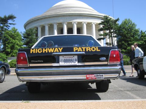 1991 Ford LTD Crown Victoria - rear of a black Ford Crown Victoria Highway Patrol car with a California plate, people standing beside it near a domed memorial.