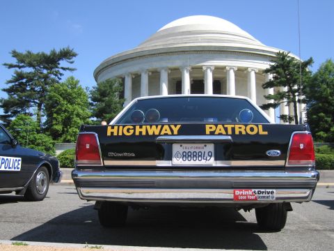 1991 Ford LTD Crown Victoria - rear view of a black Ford Crown Victoria Highway Patrol car with twin roof lights, parked before a white domed memorial.