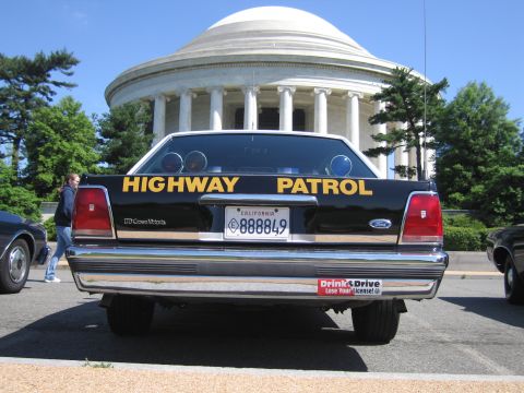 1991 Ford LTD Crown Victoria - rear of a black Ford Crown Victoria Highway Patrol car with California plate, a person in jeans walking past at a domed memorial.