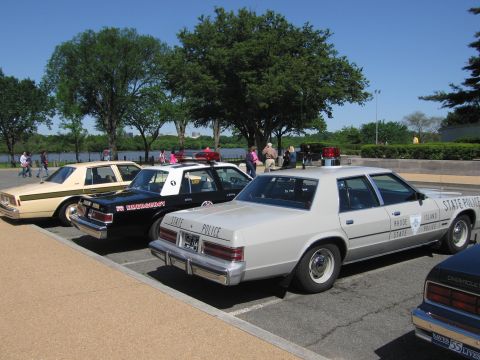 1991 Ford LTD Crown Victoria - row of police sedans angle-parked by a waterfront, led by a silver State Police car with roof beacon and a black-and-white unit behind.