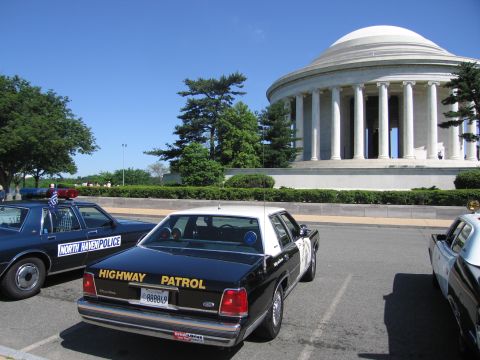 1991 Ford LTD Crown Victoria - black-and-white Highway Patrol sedan with roof spotlight parked in a lot before a white columned domed memorial under blue sky.