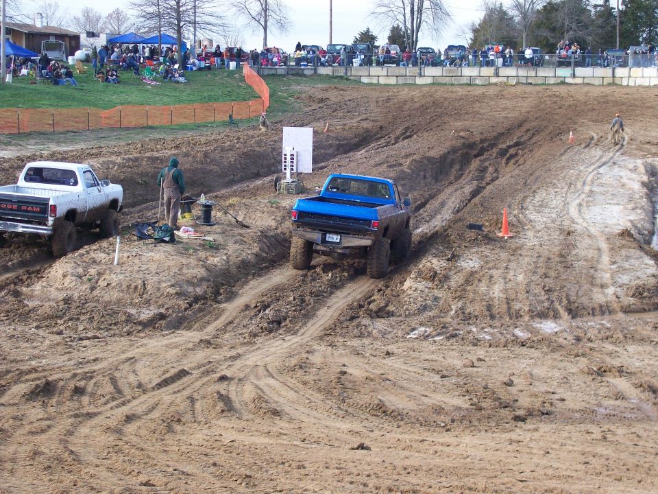 Blue pickup truck climbing a muddy hill with a white pickup truck nearby