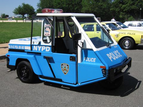 1991 Ford LTD Crown Victoria - blue-and-white NYPD three-wheeled police scooter parked beside a yellow State Police sedan on a paved lot.