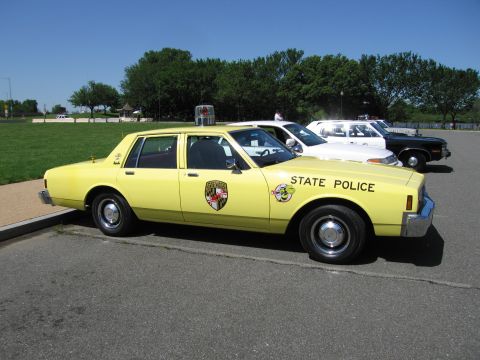 1991 Ford LTD Crown Victoria - yellow State Police sedan with a colorful door shield parked on pavement among other vehicles on a sunny day.