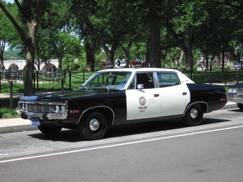 1991 Ford LTD Crown Victoria - black-and-white police sedan marked POLICE with a roof beacon parked on a road in a green park near a stone bridge.