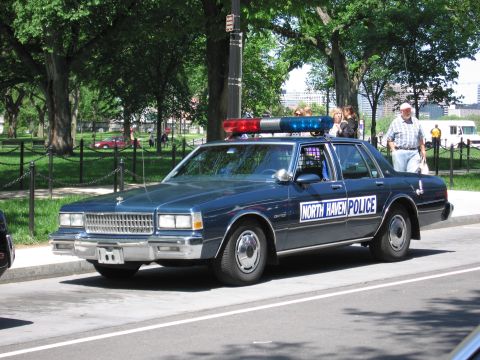 1991 Ford LTD Crown Victoria - dark blue North Haven Police Chevrolet sedan with red and blue roof lightbar parked along a road in a tree-lined park.