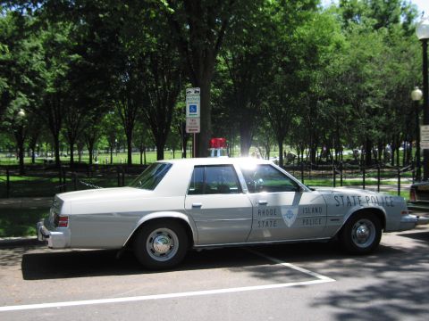 1991 Ford LTD Crown Victoria - white Rhode Island State Police four-door sedan with a single red roof beacon parked in a shaded, tree-lined parking area.