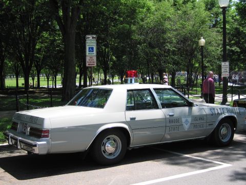 1991 Ford LTD Crown Victoria - Rear three-quarter view of a white Rhode Island State Police sedan with red roof beacon parked among trees with people in the background.