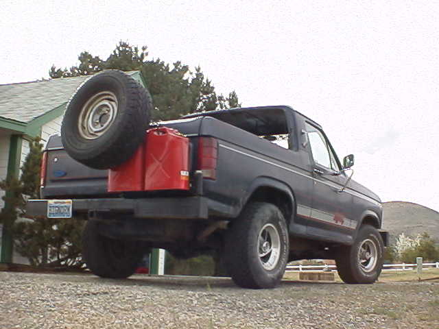 Dark blue Ford Bronco, rear 3/4 view, tailgate-mounted spare tire and red gas can, top removed