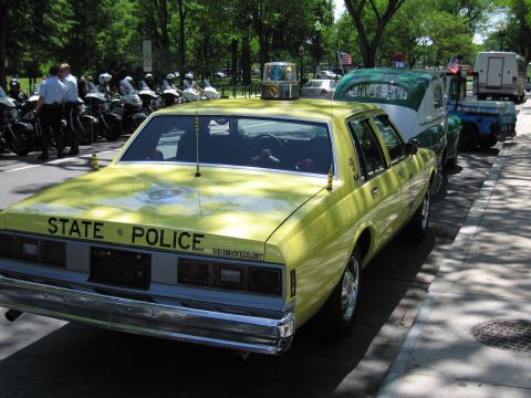 1991 Ford LTD Crown Victoria - Rear three-quarter view of a yellow Chevrolet sedan lettered STATE POLICE parked in a row with police motorcycles and other vehicles.