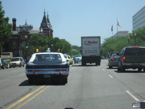 1991 Ford LTD Crown Victoria - View through a windshield of a dark vintage sedan with roof beacon driving a city street past a Ryder box truck near a Gothic-spired buildi…