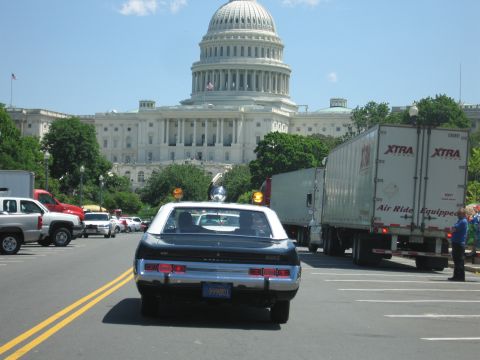 1991 Ford LTD Crown Victoria - dark vintage sedan with a roof beacon drives down a street toward the U.S. Capitol dome, passing tractor-trailers along the way.