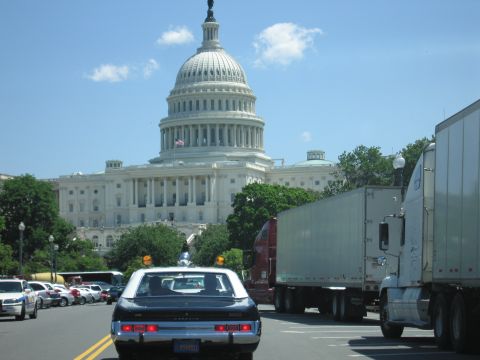 1991 Ford LTD Crown Victoria - dark vintage sedan with roof beacon drives a sunlit street with the U.S. Capitol dome and a white semi-trailer in the background.