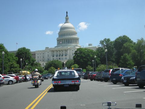 1991 Ford LTD Crown Victoria - dark vintage sedan with roof beacon and a police motorcycle drive up a parking-lined street toward the domed U.S. Capitol.