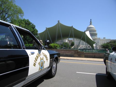 1991 Ford LTD Crown Victoria - black-and-white Highway Patrol sedan with star door emblem parked along a road near a large tent canopy and the U.S. Capitol dome.