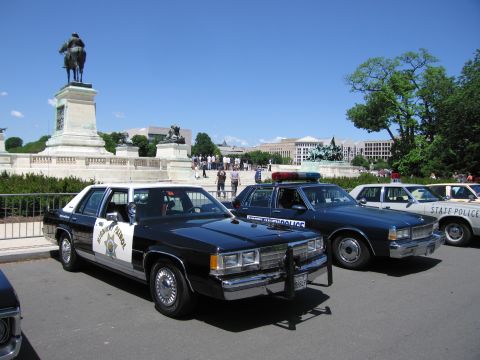 1991 Ford LTD Crown Victoria - black-and-white California Highway Patrol sedan parked among other vintage police cars near an equestrian statue and stone monument.