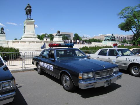 1991 Ford LTD Crown Victoria - dark blue police sedan marked POLICE parked behind a barricade near a bronze monument, with other cruisers alongside.